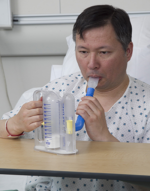Deep breathing expands the lungs, aids circulation, and helps prevent pneumonia. Man in hospital bed using incentive spirometer.